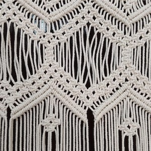 Pode incluir: Close-up de uma tapeçaria de macramé creme. O têxtil feito à mão apresenta um padrão geométrico de diamantes e chevrons, com franjas verticais na parte inferior. O design é intrincado e detalhado, mostrando os nós e a tecelagem.