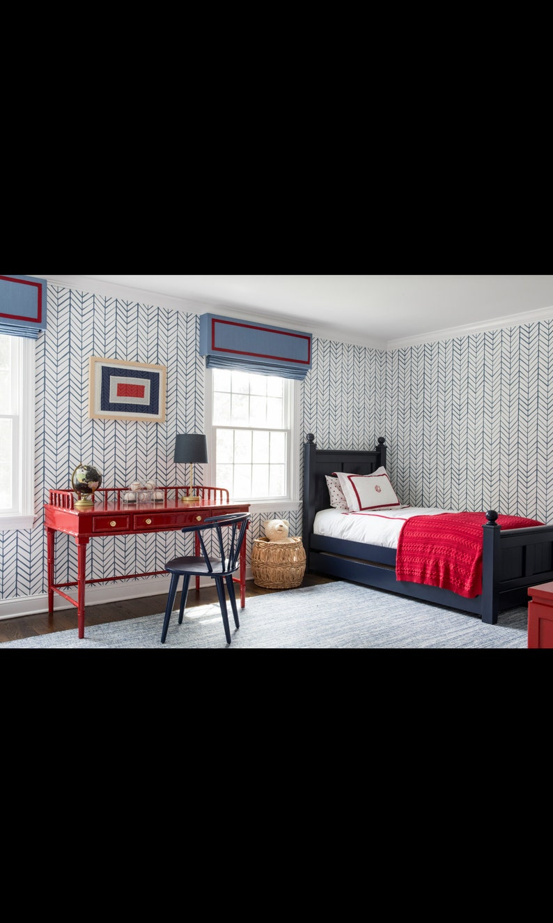 May include: A red wooden desk with drawers and a black chair sits in a room with blue and white chevron patterned wallpaper. A blue bed with a red blanket is in the background.