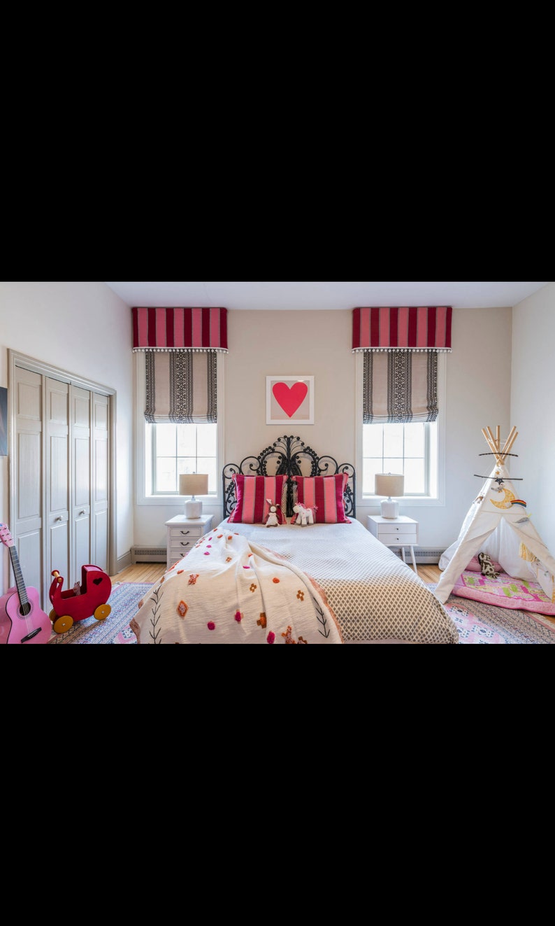 May include: A white bed with a black metal headboard and pink and white striped pillows. A white blanket with pink and orange embroidery is on the bed. There is a red toy wagon and a guitar on the floor. A teepee tent is in the corner of the room.