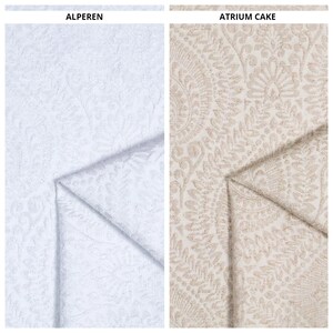 Op de afbeelding: Twee stofstalen, een wit met een subtiel reliëfpatroon en een beige met een prominenter reliëfbloemmotief. De stalen zijn gelabeld "ALPEREN" en "ATRIUM CAKE".