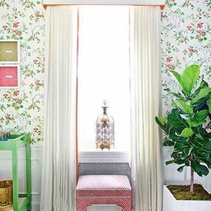 May include: A pink upholstered stool with a white base sits in front of a window with white curtains and a floral patterned wallpaper. A decorative metal lantern sits on the windowsill.