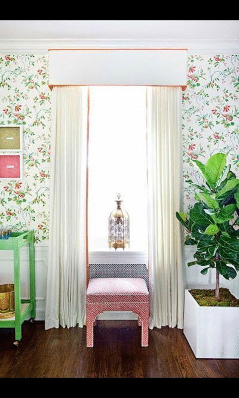 May include: A pink upholstered stool with a white base sits in front of a window with white curtains and a floral patterned wallpaper. A decorative metal lantern sits on the windowsill.