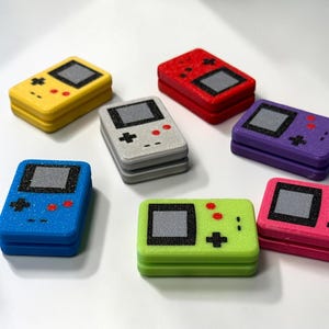 May include: A collection of seven handheld gaming console replicas in yellow, red, grey, purple, blue, lime green, and pink. Each rectangular unit features a black screen area, a directional pad, and two red circular buttons on the front surface.