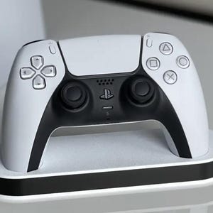 May include: A white PlayStation 5 DualSense wireless controller is sitting on a white charging dock. The controller has black accents and buttons.