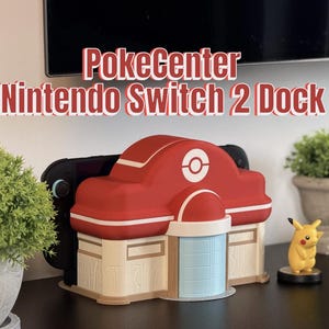 May include: A red and white PokeCenter Nintendo Switch 2 Dock. The dock is designed to resemble a building with a red roof, white accents, and a blue and beige facade. A black Nintendo Switch console is inserted into the dock. A Pikachu figurine is on the right.