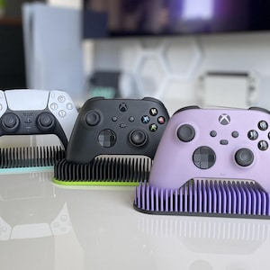 May include: Three different colored Xbox controllers on stands. The controllers are white, black, and purple. The stands are all black with a textured surface.