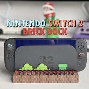 May include: A Nintendo Switch 2 Brick Dock in gray, with a pixelated landscape design. The dock holds the Switch console and game cartridges. The text "NINTENDO SWITCH 2 BRICK DOCK" is displayed in the image.