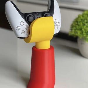 May include: A yellow and red 3D printed stand for a white and black video game controller. The stand is shaped like a cartoon character.