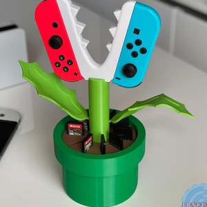 May include: A green plant-shaped Nintendo Switch game cartridge holder with a red and white controller on one side and a blue controller on the other side. The plant has a brown pot with a green base.