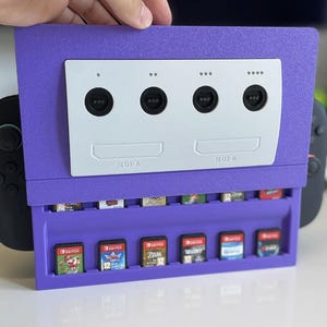 May include: A purple video game console organizer with a white faceplate and black controller ports. The organizer has a slot for game cartridges. The text "SLOT A" and "SLOT B" are visible. Several game cartridges are in the organizer.