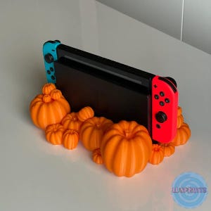 May include: A Nintendo Switch console with blue and red controllers is displayed on a stand made of orange pumpkins. The stand is shaped like a pumpkin and has a variety of sizes of pumpkins.