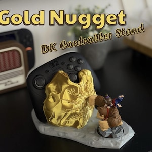 May include: A gold-colored controller stand with the text "Gold Nugget DK Controller Stand". The stand holds a black Nintendo Switch controller and features a gold nugget design and a Donkey Kong figurine.