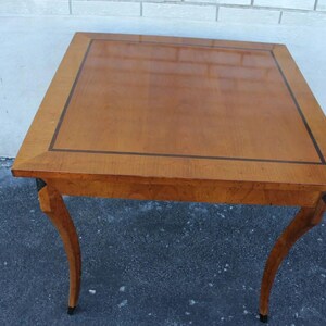 May include: A square wooden side table with a glossy, light brown surface and a dark brown border. The table has four curved legs. The table is approximately 24 inches wide and 24 inches long.