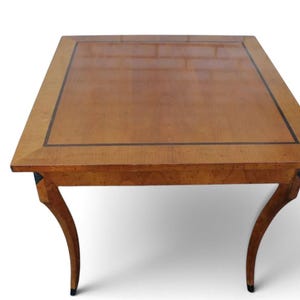 May include: A rectangular wooden table with a polished light brown finish, featuring a dark brown inlaid border on the tabletop surface and four curved legs that taper toward the bottom with dark accents at the feet and top joints.