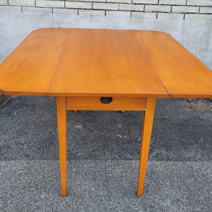 May include: A light brown wooden drop-leaf table featuring a rectangular top with rounded corners and two hinged side leaves. The table includes a single central drawer equipped with a dark metal pull handle and is supported by four straight legs.