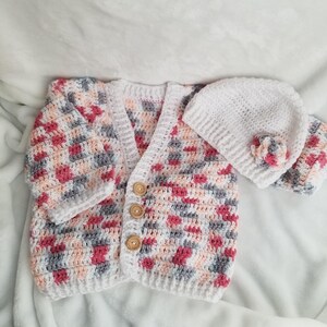May include: A crocheted baby cardigan and hat set in pink, gray, and white with wooden buttons.