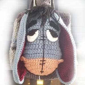 May include: A handmade crochet backpack featuring a depiction of Eeyore from Winnie the Pooh. The backpack has a gray body, blue ears, and a tan muzzle. The Eeyore face is centered on the front of the bag, with black and white details.