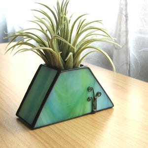 May include: A triangular stained glass planter in shades of green and blue, with a black border. It holds a green air plant. The planter is on a wooden surface, with a decorative metal accent on the side.