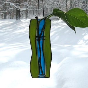 May include: A stained glass vase with a blue and green design hangs from a string. A single stem with leaves is in the vase.