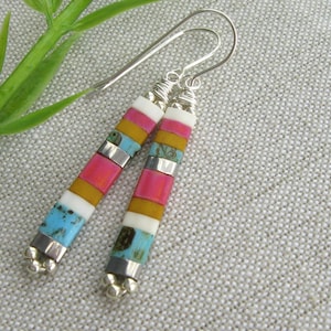 May include: A pair of dangle earrings with a colorful geometric design. The earrings are made of turquoise, pink, white, and yellow stones, and are set in silver.