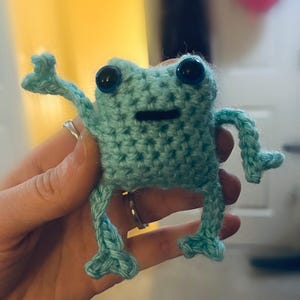 May include: A small, crocheted frog in a light teal color with large, black button eyes. The frog has outstretched arms and legs, giving it a playful appearance. The handmade amphibian is a whimsical, decorative item.