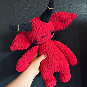 Pode incluir: Um brinquedo de pelúcia vermelho de crochê com chifres pretos e cauda. O brinquedo tem olhos pretos grandes e uma pequena boca preta.