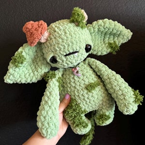 Peut inclure: Un jouet en peluche crocheté vert avec un champignon sur la tête et un petit charme de fleur autour du cou. Le jouet a de grands yeux et une petite bouche.