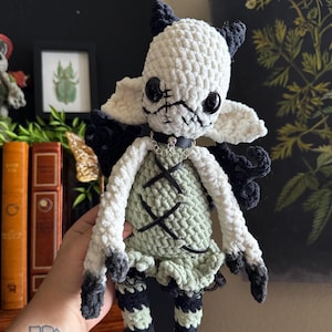 May include: A handmade crochet doll featuring a white head with black horns, large black eyes, and stitched mouth details. It wears a sage green dress with black ribbon lacing, a black collar with a silver charm, and black and white striped legs.
