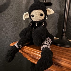 Op de afbeelding: Handgemaakte gehaakte pop met een zwarte puntmuts en bijpassend zwart lichaam. De pop heeft een lichtgekleurd gezicht, grote zwarte ogen en kleine witte oren. De benen hebben een zwart-wit patroon. Zilveren bedels hangen aan de oren.