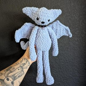 Puede incluir: Muñeca de murciélago de ganchillo azul claro con orejas y alas grandes. La muñeca tiene un collar negro con una gema morada. El murciélago está hecho a mano y tiene un aspecto suave y texturizado. El fondo es una superficie oscura y texturizada.