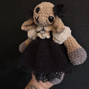 Op de afbeelding: Een gehaakt knuffeldier met beige vacht, hangende oren en zwarte knoopogen. Het draagt een zwarte jurk met een kanten zoom, een witte ruchekraag en een hartvormige hanger met een blauwe steen. Zwarte linten zijn aan het hoofd bevestigd.