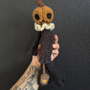 May include: A handmade crochet doll with a pumpkin head, black eyes, and a small brown stem. The doll has a white ruffled collar and a black body. A small, brown lantern hangs from the doll's legs. The doll is approximately 8 inches tall.