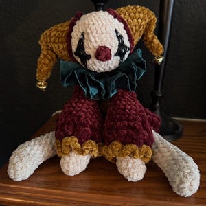 May include: A handmade plush clown doll with a cream-colored face, burgundy nose, and black eye details. The doll has a mustard yellow hat with gold bells, a green ruffled collar, and burgundy pants. The doll is sitting on a wooden surface.