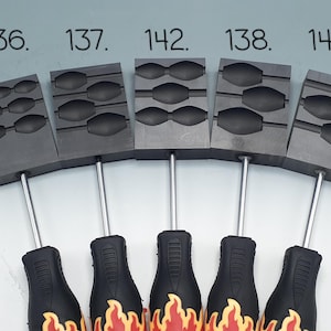 May include: Five dark grey rectangular metal casting moulds with black handles featuring a yellow and red flame graphic. Each mould has distinct cavity shapes and is labelled with the numbers 136, 137, 142, 138, and 143 printed above them.