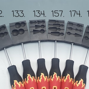 May include: Six black metal molds with different designs for casting jewelry. The molds are attached to red and black handled tools with a flame design.