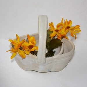 May include: A white ceramic basket with a handle, featuring a wavy design. The basket is filled with yellow artificial flowers.