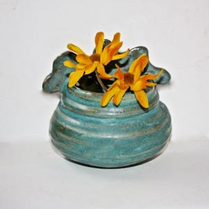 May include: A teal blue ceramic wall vase with a textured, wavy design. The vase has a rounded shape and a small opening at the top. It is decorated with two yellow flowers.