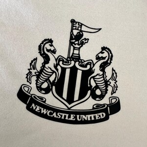 May include: Black and white crest with two seahorses, a castle, and a flag. The crest is surrounded by a banner that reads "NEWCASTLE UNITED".