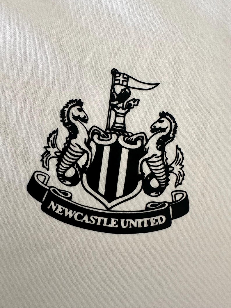 May include: Black and white crest with two seahorses, a castle, and a flag. The crest is surrounded by a banner that reads "NEWCASTLE UNITED".