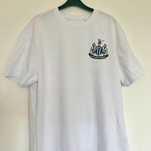 May include: White t-shirt with a black and white Newcastle United crest on the front. The crest features a shield with a black and white striped design, two black seahorses, and a black flag with a white star.