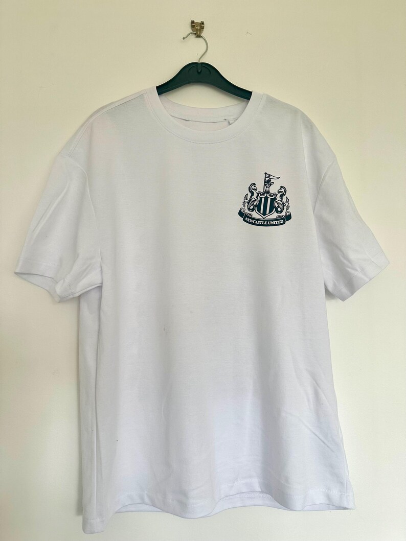 May include: White t-shirt with a black and white Newcastle United crest on the front. The crest features a shield with a black and white striped design, two black seahorses, and a black flag with a white star.