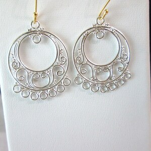 May include: A pair of silver-toned dangle earrings featuring a circular frame with intricate filigree scrollwork and a row of small open circles along the bottom edge. The earrings are attached to gold-toned metal ear wires for hanging.