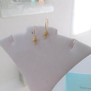May include: A pair of gold-colored dangle earrings. Each earring features a small, detailed floral design at the earlobe and a dangling element. The earrings are displayed on a white jewelry stand, showcasing their delicate design.