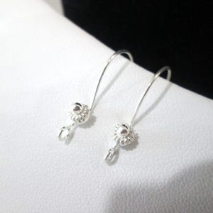 May include: A pair of silver dangle earrings. Each earring features a small, textured flower design at the top, with a teardrop-shaped charm hanging below. The earrings are displayed on a white surface.