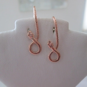 May include: A pair of rose gold snake earrings with a textured finish. The earrings are displayed on a white surface.