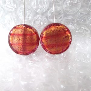 May include: A pair of round, red and gold earrings. Each earring features a transparent red glass orb with gold flecks and a silver wire for hanging. The earrings are displayed against a white, bubble-wrap background.