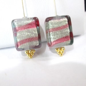 May include: A pair of drop earrings featuring rectangular glass beads with horizontal stripes of translucent gray, pink, and silver metallic foil. Each bead is accented at the bottom with a small cluster of tiny gold-toned metal spheres.