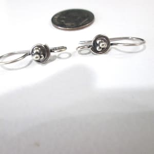 May include: A pair of silver-toned earrings. Each earring features a small, round design with a cluster of three small beads at the center. The earrings have a simple hook closure. A coin is in the background.