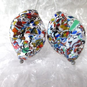 May include: A pair of colorful, teardrop-shaped earrings. The earrings feature a mosaic of red, orange, yellow, green, blue, and white glass pieces, interspersed with silver foil. Each earring is suspended from a silver wire and has a small, beaded accent at the bottom.
