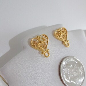 May include: Two gold-colored filigree heart-shaped charms with a small loop at the bottom. A silver dime is next to the charms. The charms are on a white surface.